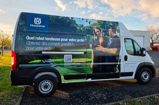 Fourgon Husqvarna Home Service garé à l’extérieur avec publicité pour un robot tondeuse, code QR et deux personnes.