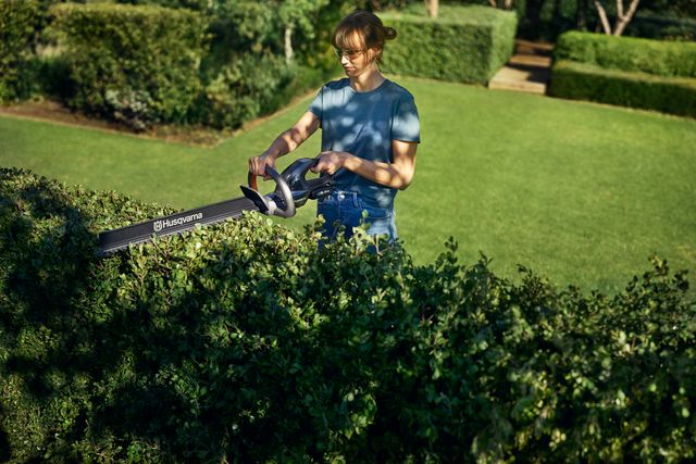 Une personne taille une haie avec un taille-haie Husqvarna dans un jardin.