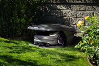 Robotic lawn mower parked in a robotic mower house in a garden near a stone wall.