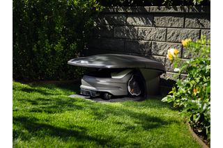 Robotic lawn mower parked in a robotic mower house in a garden near a stone wall.