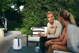 Zwei Personen sitzen auf einer Terrasse mit einem Mähroboter, der im Garten arbeitet.