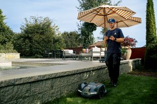 Une personne debout sur l’herbe contrôlant un robot tondeuse à l’aide d’une télécommande dans un jardin.