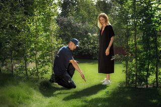 Žmogus, atsiklaupęs ant vejos sode, bendraujantis su kitu stovinčiu žmogumi.