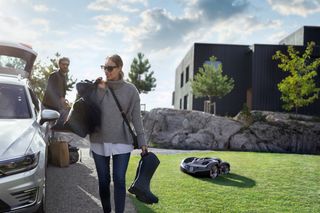 Une personne portant une paire de bottes et un sac avec un robot tondeuse fonctionnant sur une zone herbeuse à proximité.