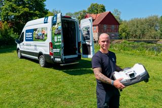 Person som håller en Husqvarna robotgräsklippare bredvid en öppen servicebil i en trädgård.