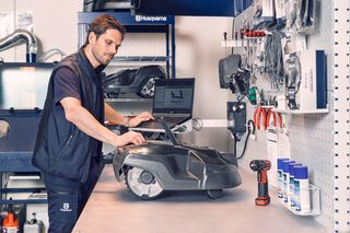 Une personne utilisant un ordinateur portable dans un atelier avec un robot tondeuse et divers outils sur la table.