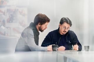 Zwei Personen sitzen mit Kaffeetassen an einem Tisch, diskutieren und schreiben Notizen.